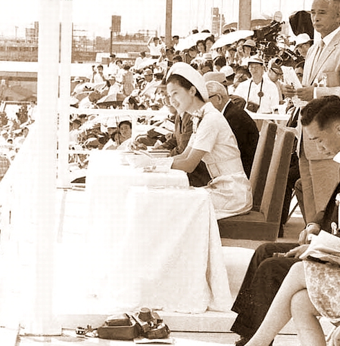 1964 10　水泳競技大会をご台覧の皇太子同妃両殿下。妃殿下の御席の左横には無造作に取材陣のカメラが。貴賓席で足を組む婦人にも注目